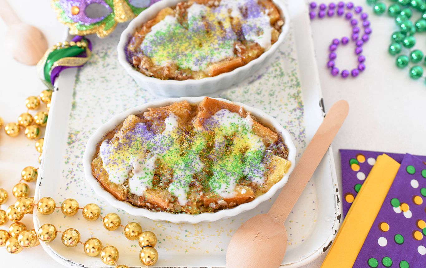 Bread pudding in white individual serving dishes topped with cream cheese frosting colored yellow purple and green on a tablescape of Mardi Gras beads